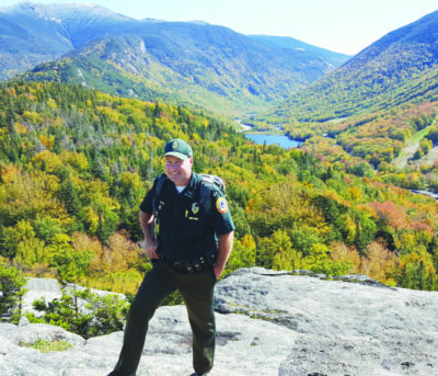 man in forestry uniform wearing baseball cap, standing on rocks in front of view of mountains in New Hampshire