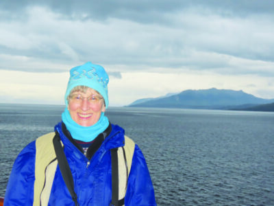 woman wearing winter coat and hat standing in front of large lake, hill in the background across the water, cloudy gray chilly looking day