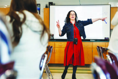 woman wearing red dress, black leggings and jean jacket standing at front of classroom pointing to white board,