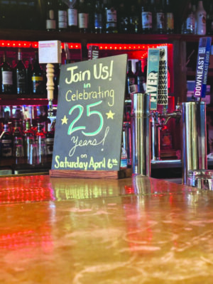 chalkboard sign on table, reading join us in celebrating 25 years, bottle of liquor in background