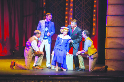 Scene from stage musical with woman wearing 1940's style dress, sitting on suitcase, surrounded by 4 men singing to her.