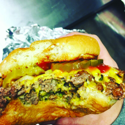 hand holding burger in foil with melty cheese ketchup and pickles, bite taken out