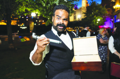black man with beard holding an open box in one hand and a pipe in the other, dressed in evening wear, outside at night on sidewalk