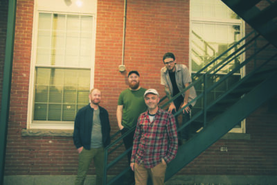 4 male band members posing outside of brick building, standing on stairway
