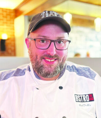 headshot of man with beard wearing glasses and baseball cap, in chef's jacket