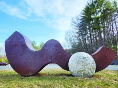 outdoor sculpture on grass on sunny day, sculpture composed of large abstract metal piece like squiggle on ground, beside stone ball