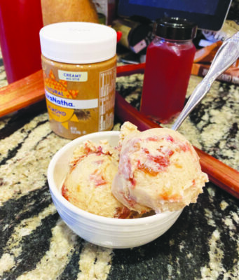 cup of ice cream with 2 scoops, on table beside jar of almond butter and bottle of rhubarb syrup sitting on counter