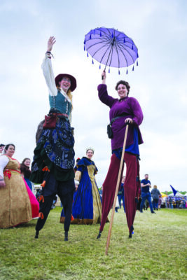 two people on stilts dressed in medieval fantasy costumes, one holding up tasseled umbrella 