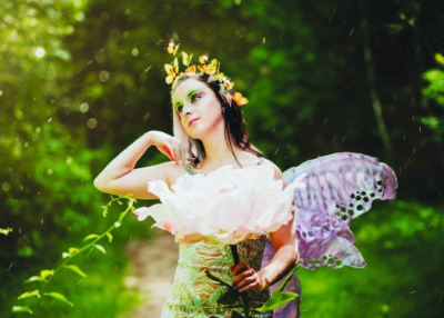 woman dressed as a fairy with wings, butterflies around her head, holding a large rose, in woods in sunlight