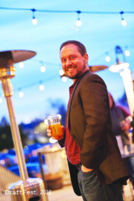 man standing half turned toward camera, holding glass of beer, one hand in pocket, wearing blazer, dusk with string lights above