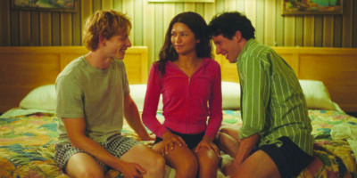 Mike Faist, Zendaya, and Josh O'Connor in a scene from Challengers - all three sitting on a bed, turned toward each other, smiling at each other