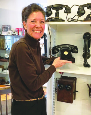 woman with brown hair wearing a black turtleneck holding a vintage telephone and smiling at the camera