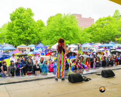 person with long hair tied with big rainbow bow, wearing rainbow striped overalls, standing on outdoor stage speaking to audience standing in front, rainy day, people holding umbrellas
