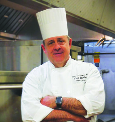 man in white chef's jacket and hat, standing in kitchen smiling with arms crossed