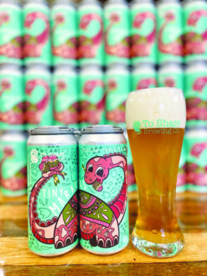 cans of beer illustrated with dinosaur playing maracas beside tall glass of beer with foam on top, in front of stacks of beer cans out of focus in background