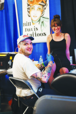 woman sitting on table while man wearing baseball cap tattoos her leg at indoor expo