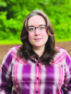 headshot of young woman wearing glasses and plaid shirt standing outside, small smile