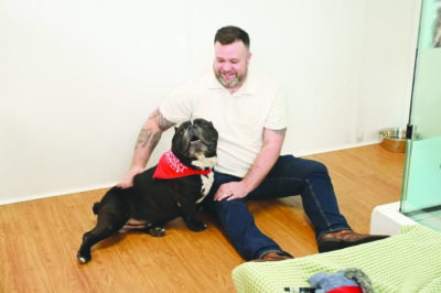 man sitting on floor dog wearing red bandana, man smiling at dog and dog cuddling up to man's side