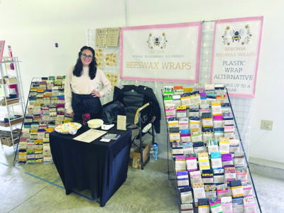 young woman wearing glasses, standing beside racks of packaged beeswax wraps with brand sign hanging on wall behind her