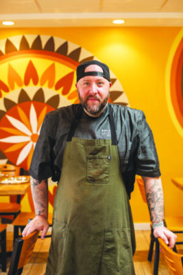 man standing in warm toned restaurant with circular design on wall, wearing backwards baseball cap and apron