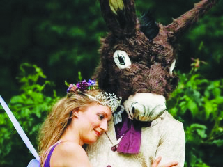 characters from Shakespeare play A Midsummer Night's Dream in costume as a female fairy with a crown and flowers in her hair embracing a man with a donkey's head