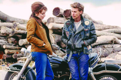 actors Jodie Comer and Austin Butler in scene from The Bikeriders standing outside talking, Butler leaning against motorcycles