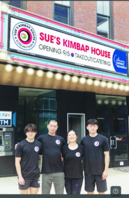 Korean family of woman, man and two boys standing in front of restaurant front with big sign reading Sue's Kimbap House