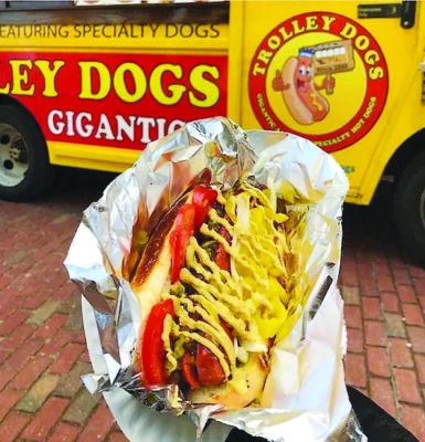 hotdog with lots of fixings in foil, being held up in front of yellow food truck with Trolley Dogs logo painted on door