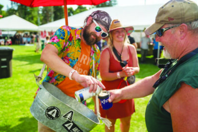 man carrying around a metal bucket, pouring another man a cup of beer from can, on sunny day during festival outside of event tents