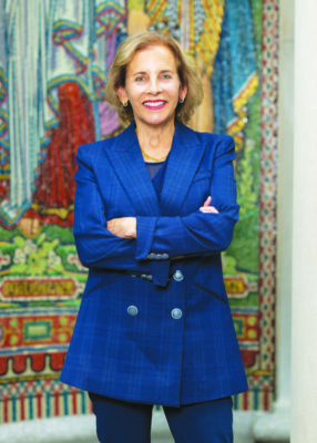 middle aged woman posing in front of art tapestry, arms crossed over chest, wearing long blue blazer, smiling