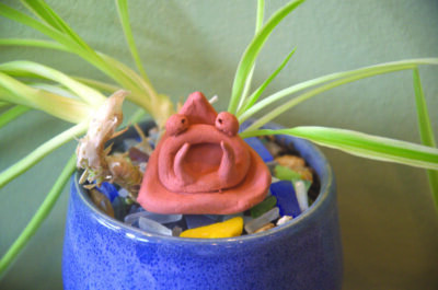 small triangular shaped monster with large mouth and tusks, big eyes, made out of red clay, sitting in potted plant