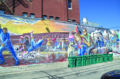 mural on long side of brick building showing scenes of people living in city, row of trash bins set out on one end