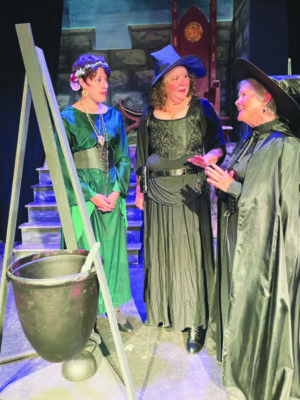 3 women dressed in long gowns standing beside prop cauldron on stage during performance