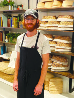 man with beard, wearing glasses and baseball cap, black apron over t-shirt, standing in front of shelves with overturned bread baskets in bakery