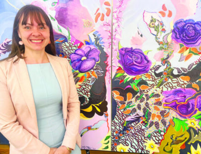 woman wearing light pink blazer standing in front of large painting of colorful flowers, smiling