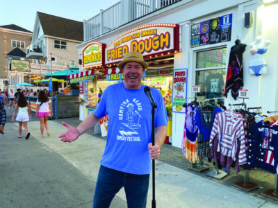 man wearing boater hat and blue t-shirt, standing on street with standing microphone at dusk