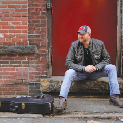 man sitting on sidewalk outside brick wall guitar at his feet