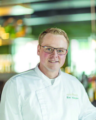 headshot of young man with short hair and round face, wearing glasses and white chef's jacket, blurry background