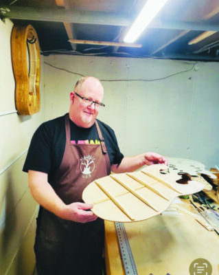 man standing in workshop holding wooden guitar face that he is making