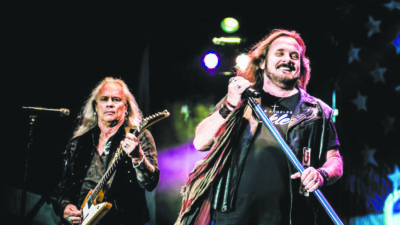 2 long haired older men on dark stage, one with guitar and one with standing microphone, during performance