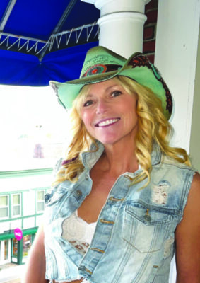 blond woman with long hair, wearing denim vest and cowboy hat smiling near window