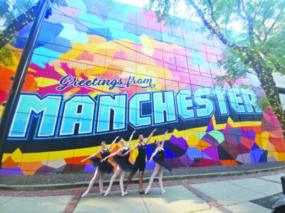 Ballet dancers posing in front of mural accross from Palace Theather that reads "Greetings from Manchester"