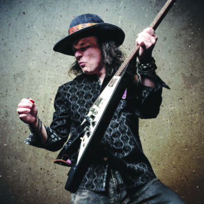 elderly man with long hair, wearing hat with brim, holding up guitar as if playing energetically