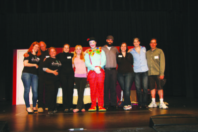 group of people, one dressed as a clown, standing in line across stage, arms around each other, dark curtain in background