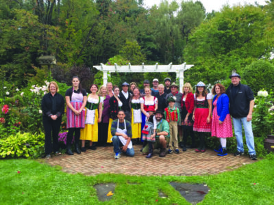 large group of people, some of them in Oktoberfest costumes standing in front of white trellis on outdoor patio, tree in background, posing for photo