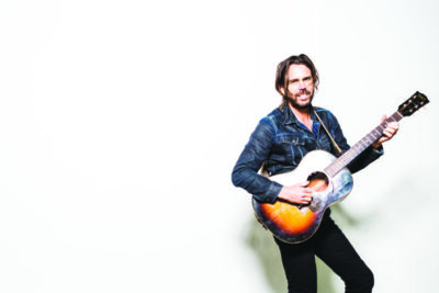 man with long hair tied up, beard and mustache, wearing denim jacket, holding guitar in playing position, posing against white wall