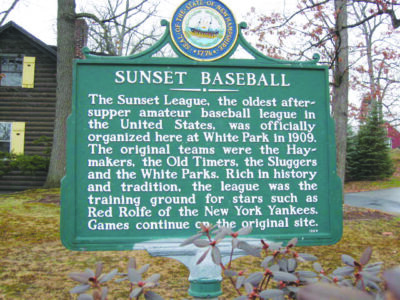 NH historical marker, green sign with white text, topped by NH state emblem, in front of old building on gray autumn day