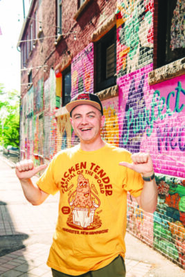 man wearing yellow shirt and baseball cap, standing in alley in front of colorful mural painted brick wall, pointing to himself with both thumbs, big smile