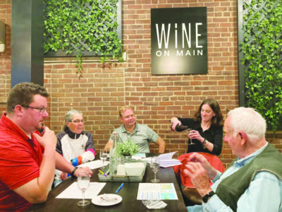 people sitting at long table in front of brick wall, paper sheets in front of them, one woman pouring wine