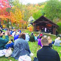 people sitting on grassy hill on autumn day, watching performance on enclosed stage at bottom of the hill